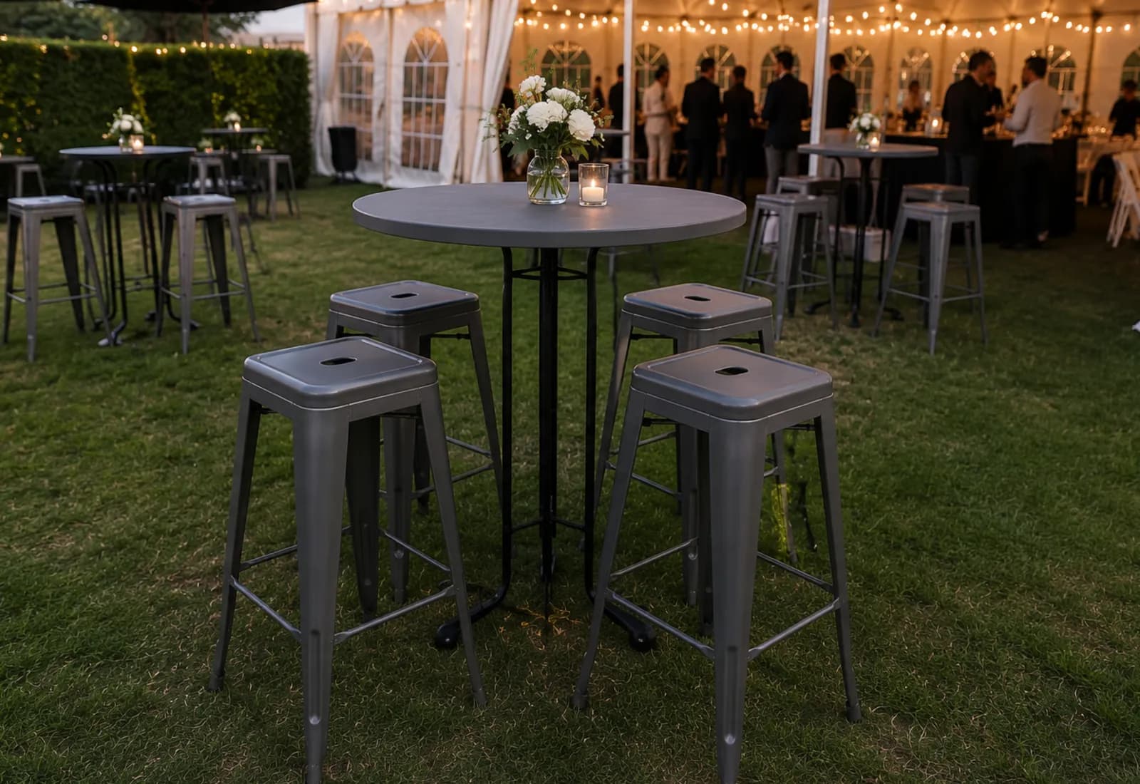 Grey round bar tables on a lawn beside a marquee at dusk